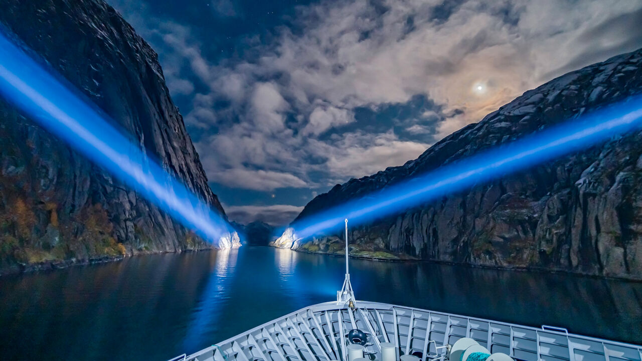Ein Kreuzfahrtschiff beleuchtet nachts mit Scheinwerfern die Felswände des Trollfjords. 