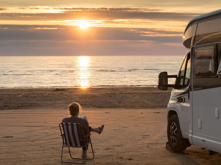 Frau liest am Strand ein Buch, Wohnmobil steht daneben 