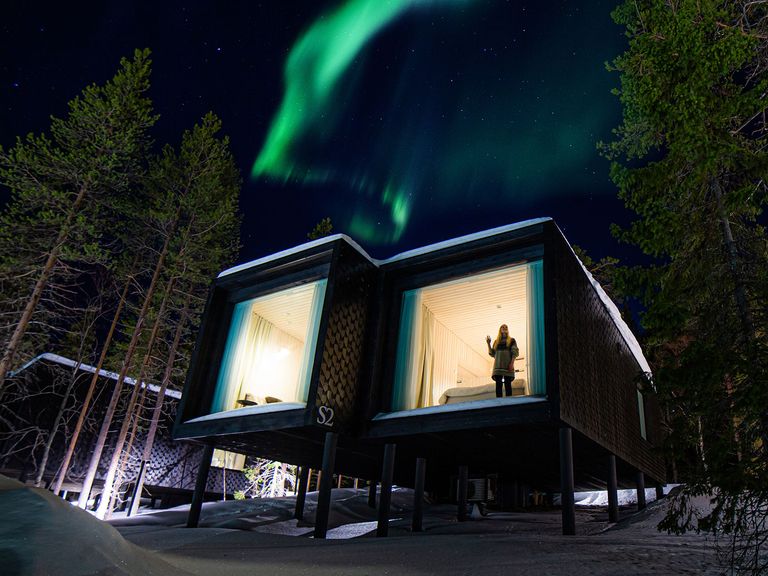 Rund um das „Arctic TreeHouse Hotel“ gibt es zahlreiche Möglichkeiten, Polarlichter zu erhaschen. Frau steht an der Scheibe eines Hotels, unterm Polarlicht