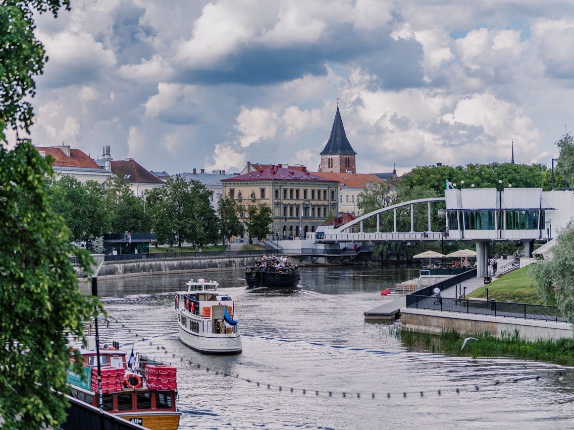 Blick auf den Fluss Emajõgi in Tartu mit vorbeifahrenden Booten, einer weißen Fußgängerbrücke und historischen Stadtgebäuden unter einem wolkenverhangenen Himmel.