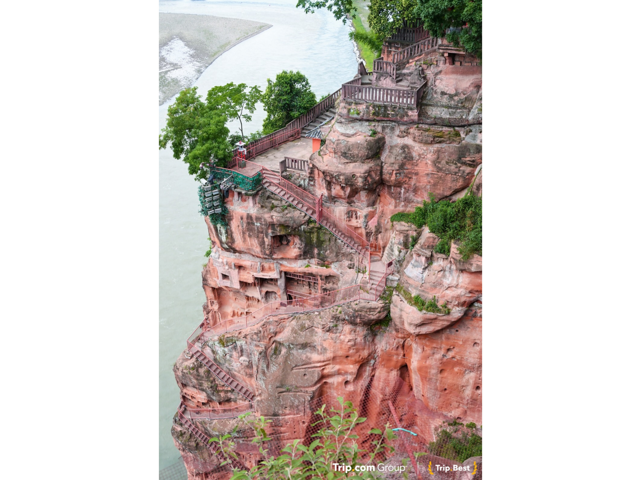 Rötliche Felswand mit in den Stein gehauenen Treppen und Pflanzenbewuchs, unten ein Fluss