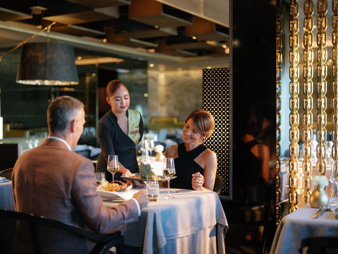 Ein Paar sitzt in einem eleganten Restaurant an Bord, während eine Servicekraft ein Gericht serviert.