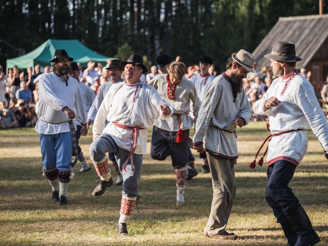 Eine Gruppe estnischer M&auml;nner in traditionellen wei&szlig;en Leinenkitteln mit roten Kordelg&uuml;rteln und H&uuml;ten vollf&uuml;hrt einen rhythmischen Volkstanz auf einer Wiese vor Publikum.