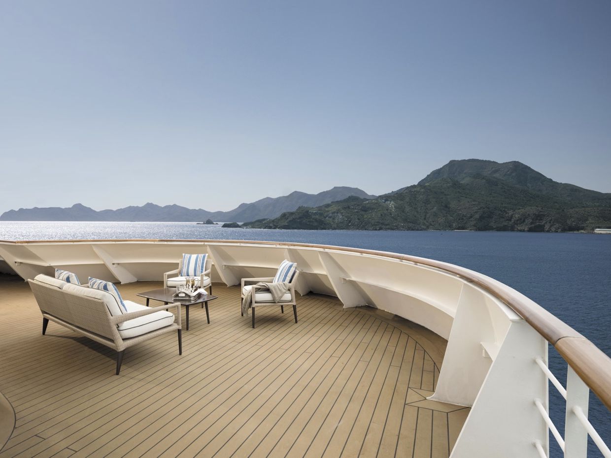 Ein weitläufiges Außendeck mit Lounge-Möbeln vor einer bergigen Küstenlandschaft und offenem Meer.