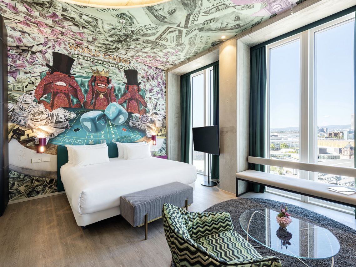 Junior Suite im nhow Frankfurt mit wei&szlig;em Bett, gro&szlig;em Wandmotiv, hohen Fenstern und Blick &uuml;ber Frankfurt.