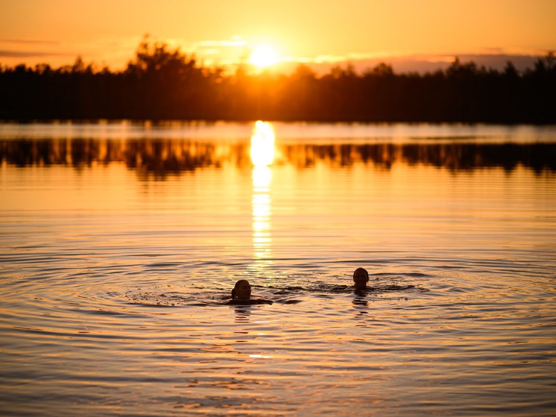 Zwei Personen schwimmen bei Sonnenuntergang in einem ruhigen Moorsee in Estland, w&auml;hrend sich das orangefarbene Licht im Wasser spiegelt.