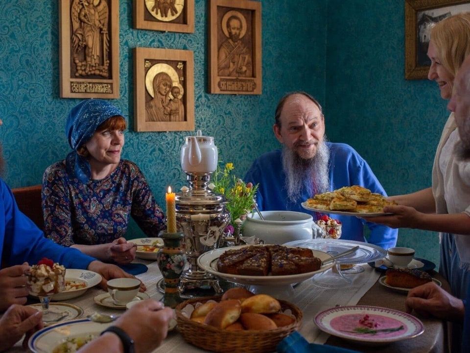 Eine Gruppe von Menschen sitzt in einem Raum mit gemusterten blauen Tapeten und religi&ouml;sen Ikonen um einen reich gedeckten Tisch mit einem gl&auml;nzenden Samowar, w&auml;hrend Kuchen serviert wird.