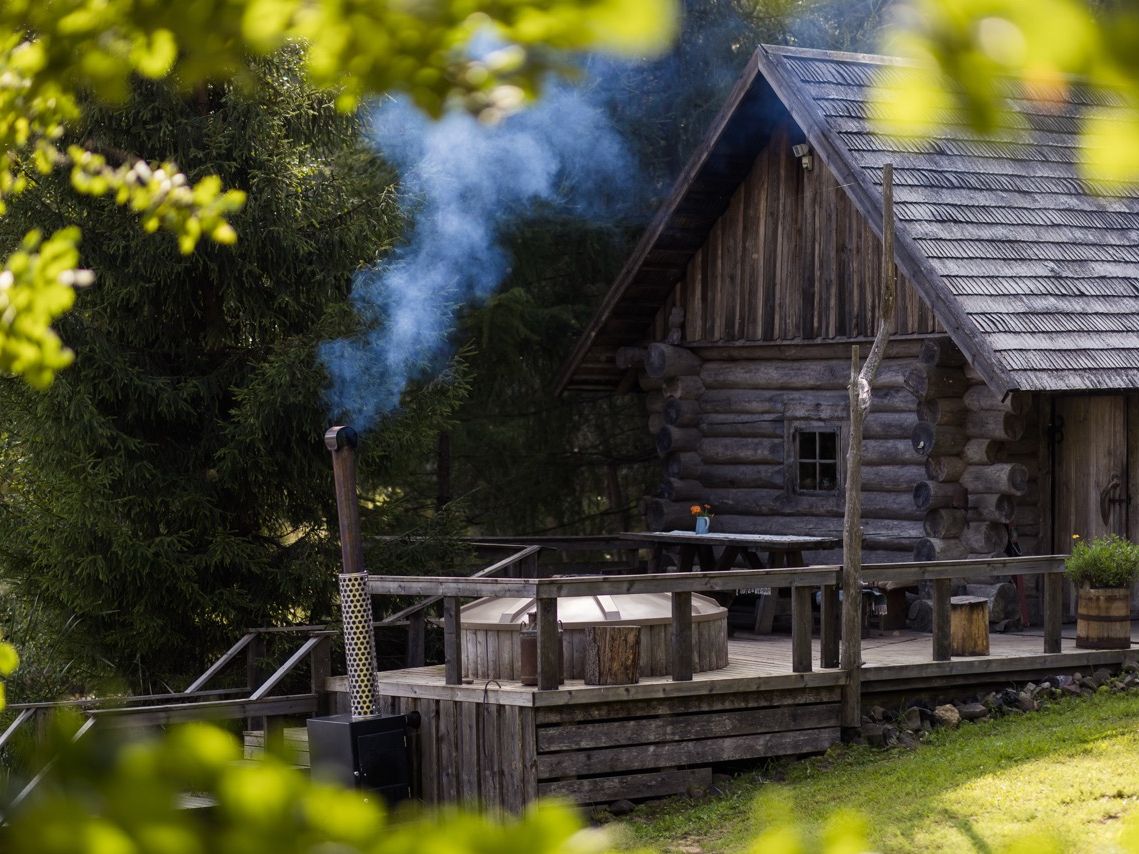 Eine urige, traditionelle Blockhaus-Rauchsauna in Estland mit einer Holzterrasse, einem Badezuber und rauchendem Schornstein vor dichten Nadelb&auml;umen.