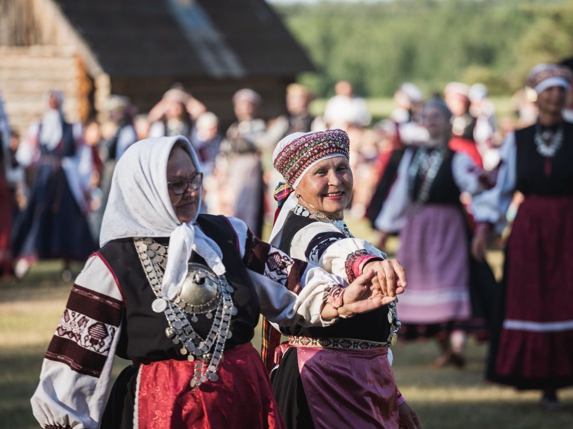 Zwei l&auml;chelnde Frauen in traditioneller estnischer Seto-Tracht mit wei&szlig;en Kopft&uuml;chern, bestickten &Auml;rmeln und auff&auml;lligem Silberschmuck tanzen Hand in Hand im Freien.