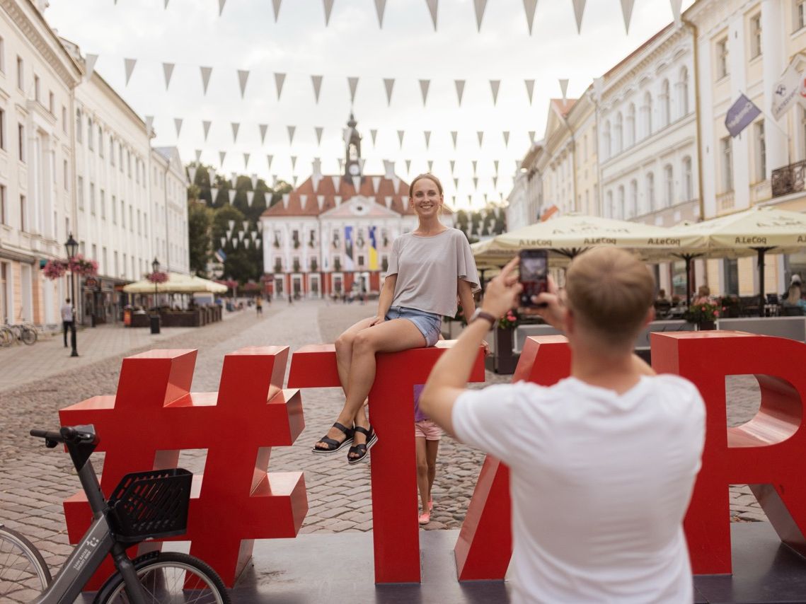 Ein Mann fotografiert eine Frau, die im Sommer auf einer gro&szlig;en, roten Buchstaben-Skulptur auf dem kopfsteingepflasterten Rathausplatz in Tartu sitzt.