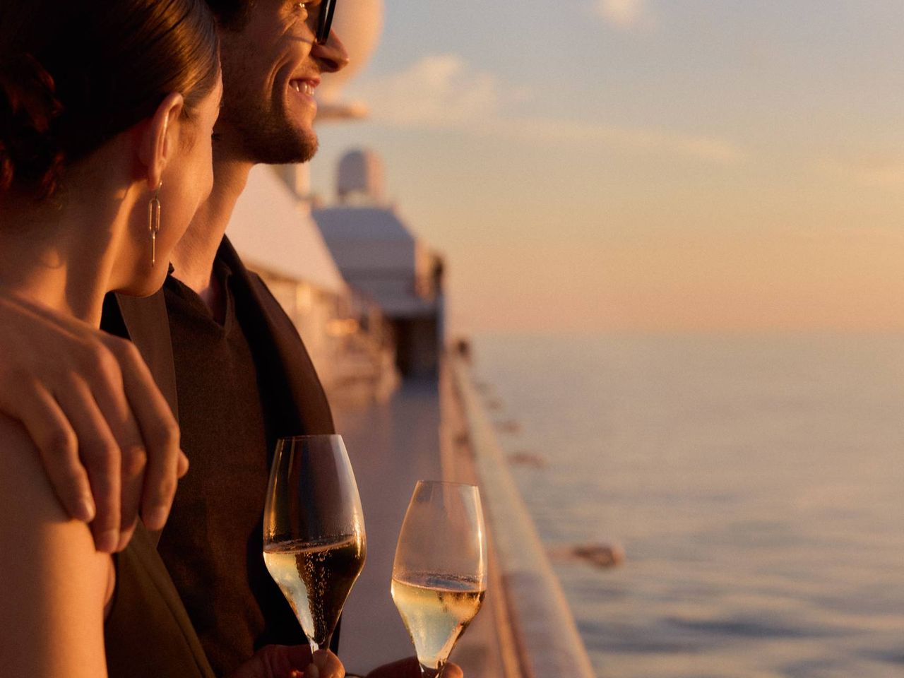 Zwei G&auml;ste mit Champagnergl&auml;sern an Deck eines Hapag-Lloyd-Cruises-Schiffs bei Sonnenuntergang.