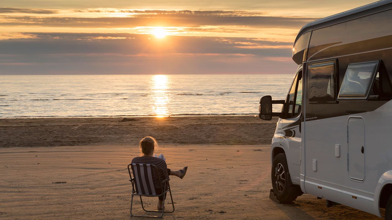 Frau liest am Strand ein Buch, Wohnmobil steht daneben 
